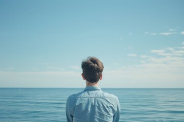 Fototapeta premium A contemplative young man gazes at the vast blue ocean, a scene of serene solitude and expansive calm, perfect for themes of reflection and nature.
