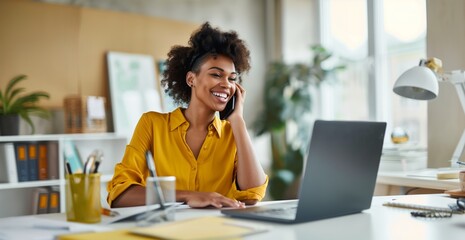 A woman is sitting at a desk with a laptop and a cell phone. She is smiling and talking on the phone,wearing yellow clothes