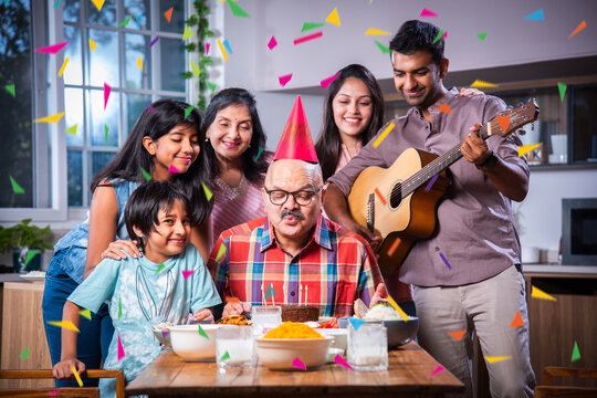 Indian asian grandparents with Family celebrating birthday by blowing candles on cake at home on dining table.