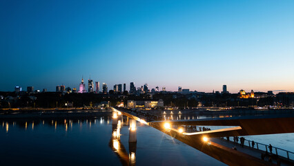 Fototapeta premium newly built pedestrian and bicycle modern bridge over Vistula river at night, illuminated contemporary architecture of Warsaw, aerial top view