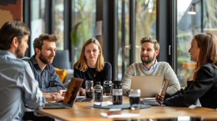A group of young people at a meeting in a modern conference room in an office. Colleagues are sitting at a large table with laptops discussing work indoors. Meetings concept. - Powered by Adobe