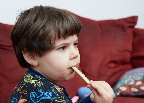 cute baby boy eating a cucumber on the couch in his dinasaur pajamas
