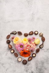 Heart made with delicious chocolate candies and beautiful flowers on grey table, flat lay. Space for text