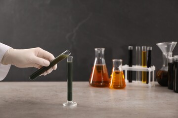 Woman pouring green crude oil into test tube at grey table, closeup
