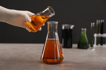 Woman pouring yellow crude oil into flask at grey table against dark background, closeup