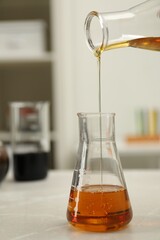 Pouring orange crude oil into flask at light marble table, closeup