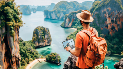 Fototapeta premium A traveler with a backpack stands on a cliff with a laptop overlooking a stunning tropical bay.