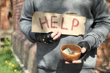 Poor homeless man holding help sign and bowl with donations outdoors, closeup