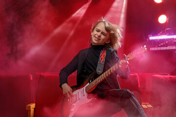 A boy plays an electric guitar in a red interior in the rays of stage spotlights.
