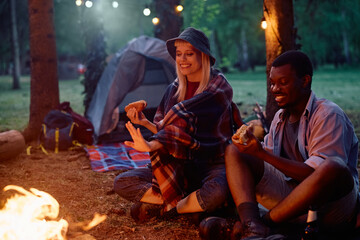 Happy couple eating hot dogs by campfire in nature.