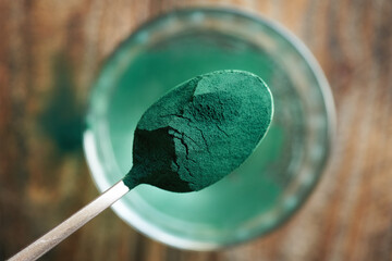 Green spirulina powder on a metal spoon above a glass of waterp