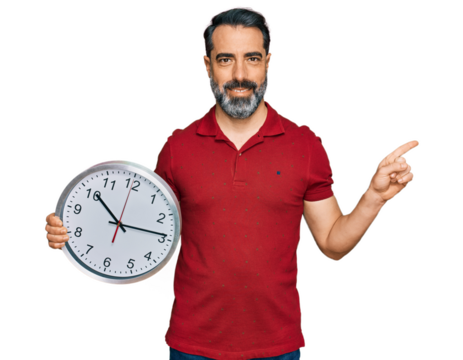 Middle aged man with beard holding big clock smiling happy pointing with hand and finger to the side