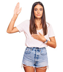 Young hispanic woman wearing casual white tshirt swearing with hand on chest and open palm, making...