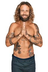 Fototapeta premium Handsome man with beard and long hair standing shirtless showing tattoos praying with hands together asking for forgiveness smiling confident.