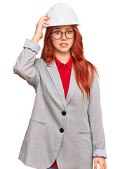 Young redhead woman wearing architect hardhat confuse and wonder about question. uncertain with doubt, thinking with hand on head. pensive concept.