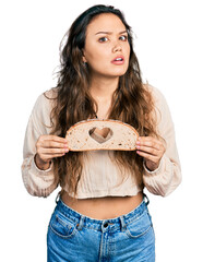 Young hispanic girl holding bread with heart symbol clueless and confused expression. doubt concept.