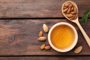Almond oil in bowl, nuts, spoon and leaves on wooden table, flat lay. Space for text