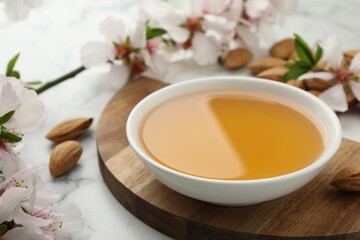 Almond oil in bowl, nuts and flowers on white marble table, closeup