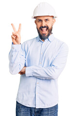 Young handsome man wearing architect hardhat smiling with happy face winking at the camera doing victory sign. number two.