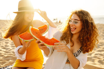 Female friends on vacation at the beach in summer eating a watermelon with the sea in the background, sitting on the sand at sunset. People, lifestyle, travel, nature and vacations concept.