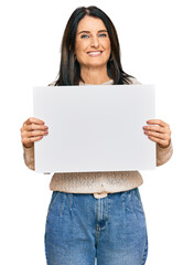 Fototapeta premium Middle age brunette woman holding blank empty banner looking positive and happy standing and smiling with a confident smile showing teeth