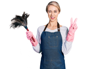Beautiful young blonde woman wearing apron holding cleaning duster smiling with happy face winking at the camera doing victory sign with fingers. number two.