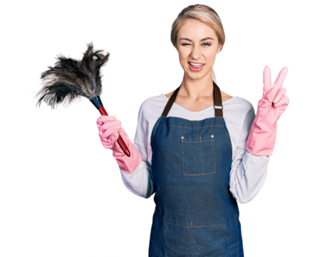 Beautiful young blonde woman wearing apron holding cleaning duster smiling with happy face winking at the camera doing victory sign with fingers. number two.