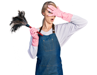 Beautiful young blonde woman wearing apron holding cleaning duster peeking in shock covering face and eyes with hand, looking through fingers with embarrassed expression.