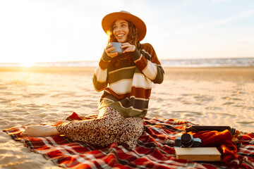 Young woman sits at picnic on the beach drinks a hot drink from a thermos. A girl enjoying beautiful view of the sea. Travel, weekend, relax and lifestyle concept