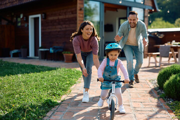 Little girl learning to ride bicycle with help of her parents in backyard.