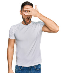 Handsome man with beard wearing casual white t shirt peeking in shock covering face and eyes with hand, looking through fingers with embarrassed expression.