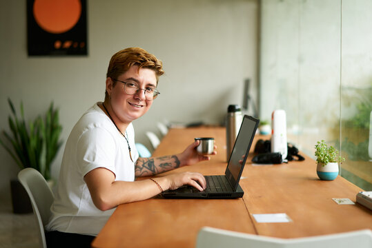 Transgender freelancer smiles at camera, works on laptop in modern office space. Sips tea from metal cup, casual work environment. Inclusive workspace promotes diversity, tech and creativity.