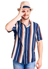 Young handsome caucasian man wearing summer hat doing ok gesture with hand smiling, eye looking through fingers with happy face.