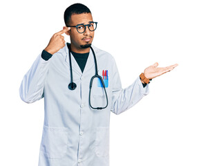 Fototapeta premium Young african american man wearing doctor uniform and stethoscope confused and annoyed with open palm showing copy space and pointing finger to forehead. think about it.