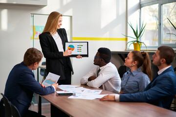 Female leader presents colorful pie chart on tablet to colleagues during office meeting. Attentive team engages in strategic planning, analysis, empowerment workplace.