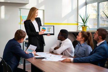 Female leader presents colorful pie chart on tablet to colleagues during office meeting. Attentive...