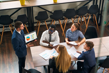 Arabic executive shares data on tablet with multiethnic team in modern office. Colleagues discuss strategy, graphs. Engaged diverse group analyzes market, plans goals at corporate meeting.
