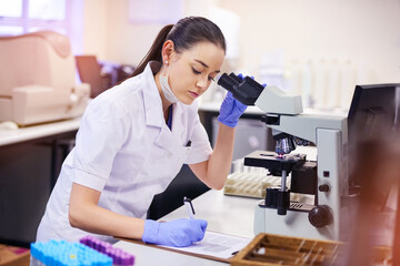 Female scientist, analyse and microscope in laboratory for research or notes with medical breakthrough. Doctor, data and health for expert in microbiology with equipment for clinical trial in London.
