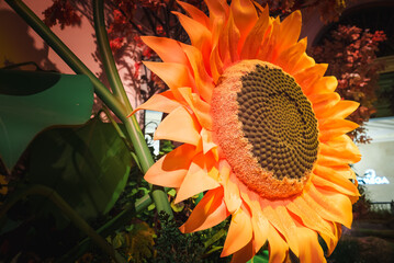 Close up view of large, vibrant artificial sunflower against green foliage backdrop, with cursive neon sign reading Flora in softly illuminated background. Location likely on Las Vegas Strip. - Powered by Adobe