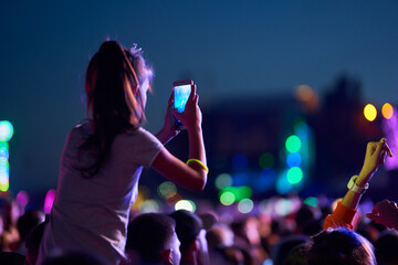 Girl films music concert on smartphone amid lively festival crowd at twilight. Audience enjoys live...