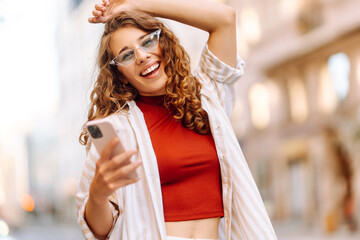 Fototapeta premium Beautiful smiling woman using phone walking in city street. Business, education, technology, blogging, communication concept.