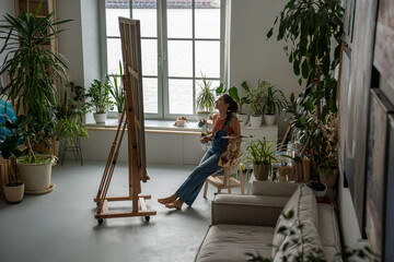 Satisfied artistic woman sitting relaxed on chair at easel holding brushes and palette with paints...