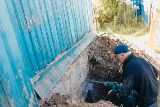 male worker drills the foundation with a hammer drill for sewerage in a country house.