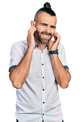 Hispanic man with ponytail wearing casual white shirt covering ears with fingers with annoyed expression for the noise of loud music. deaf concept.