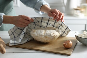 Woman covering dough with napkin at white wooden table in kitchen, closeup