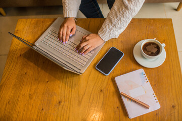 primer plano de manos de mujer escribiendo en el computador portátil con el celular y un café al...
