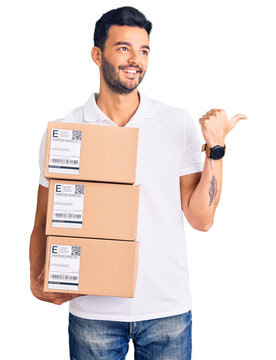 Young handsome hispanic man holding delivery package pointing thumb up to the side smiling happy with open mouth