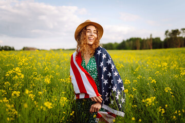 Beautiful young woman holding an American flag in the wind in a field. Summer landscape against the...