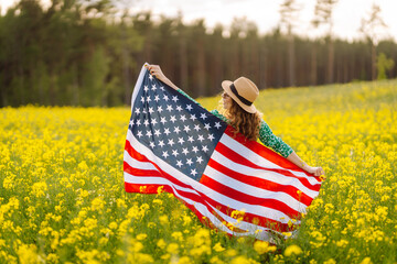 Beautiful young woman holding an American flag in the wind in a field. Summer landscape against the blue sky. Independence Day.