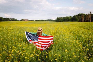 Beautiful young woman holding an American flag in the wind in a field. Summer landscape against the blue sky. Independence Day.